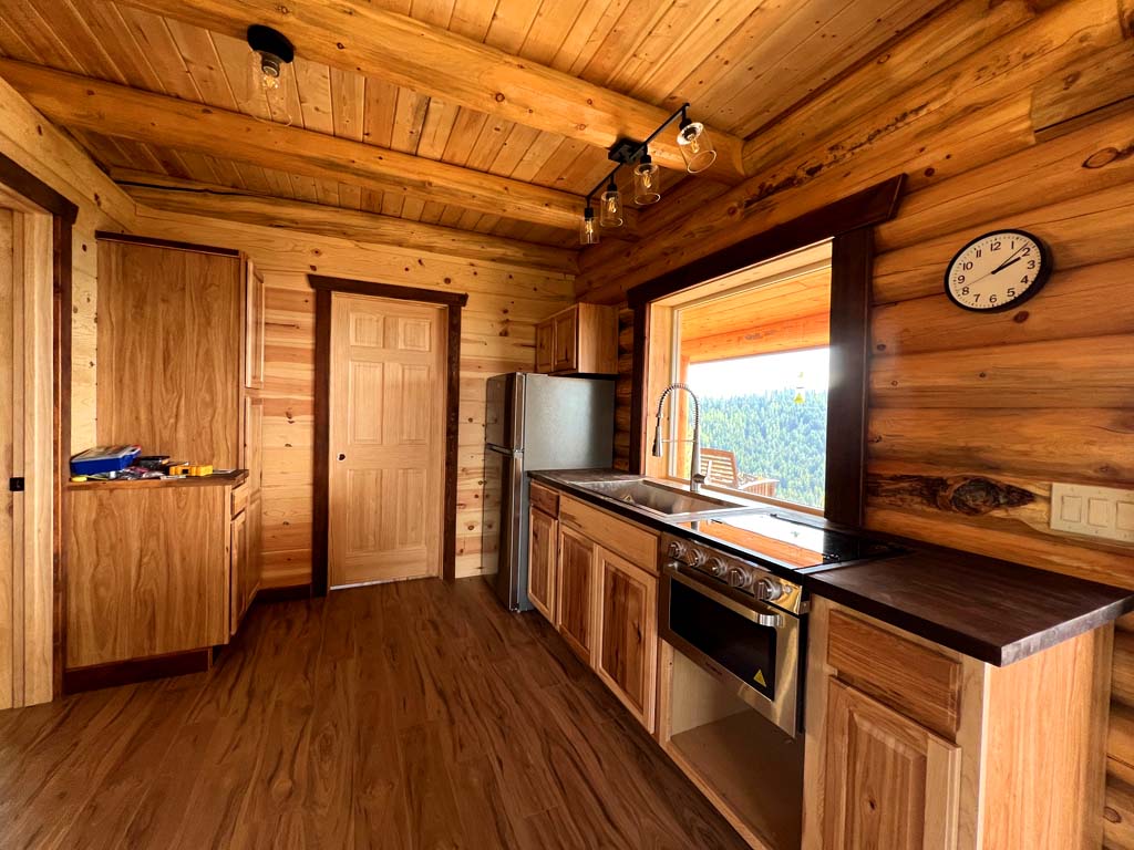 A wooden log home kitchen interior with a stainless steel refrigerator, a sink, and a wall clock. The walls and ceiling are made of rustic wood, and there is a door leading to another room.