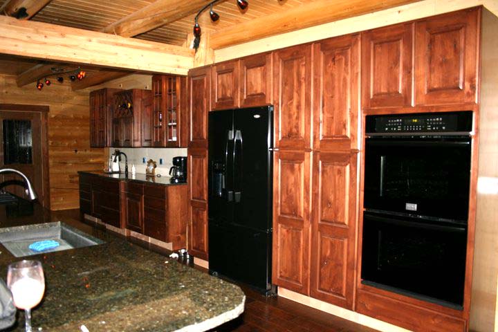 A modern log home kitchen featuring dark wooden cabinets, a black refrigerator, and built-in ovens. The countertop is made of dark stone with a glass of beverage on it. The ceiling has exposed wooden beams.
