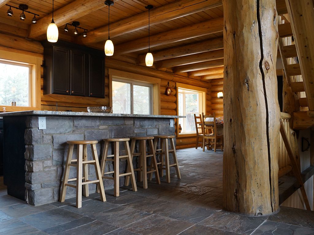 Interior view of a rustic log home kitchen with a stone island, wooden stools, wooden beams, and large windows.