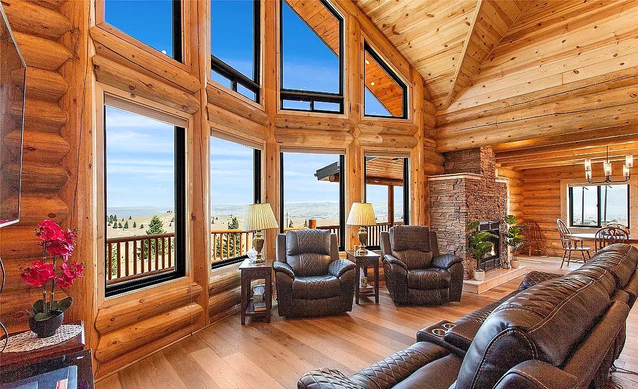 A cozy log home interior featuring large windows with mountain views, rustic wooden beams, and a stone fireplace. The room has leather recliners and a warm, inviting atmosphere.