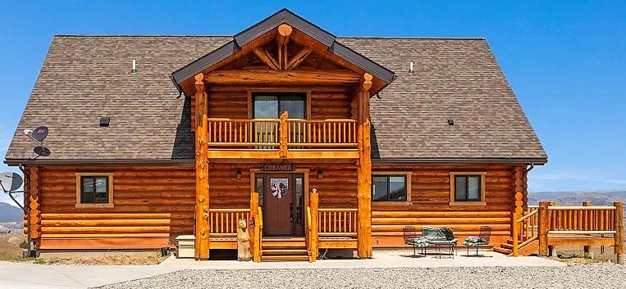 A large log cabin-style house with a peaked roof, multiple balconies, and a gravel driveway. Clear blue sky in the background.