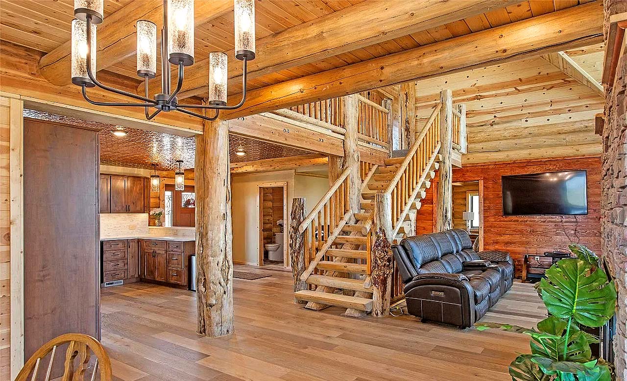 A spacious log home cabin interior featuring wooden beams, a staircase, a leather recliner, and an open kitchen area with rustic decor.