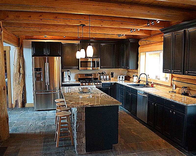 A rustic log home kitchen featuring dark cabinetry, stainless steel appliances, and a large island with a granite countertop. The ceiling has exposed wooden beams, and the floor is made of stone tiles.