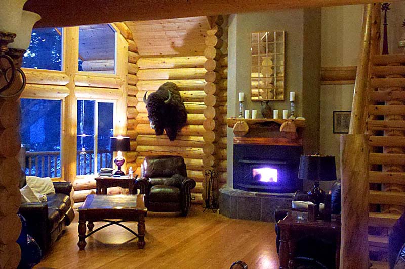 A cozy log home living room with large windows, a stone fireplace, comfortable furniture, and rustic decor.