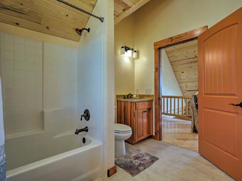 A log home bathroom featuring a shower/tub combo, toilet, and wooden accents. There's a door leading to another room visible in the background.