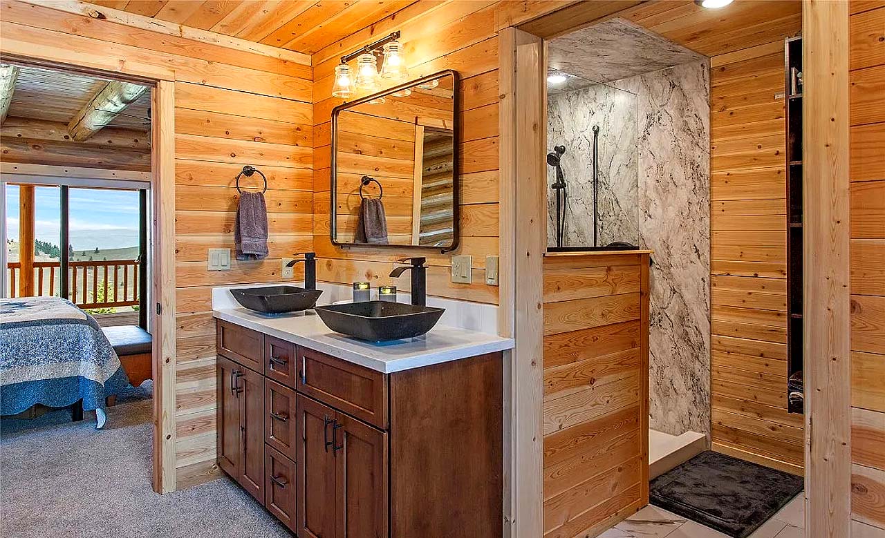 A log home wooden bathroom with dual sinks, a shower area with marble-like tiles, and a view of an outdoor space. The walls are made of horizontal wooden planks, and there are two mirrors above the sinks.