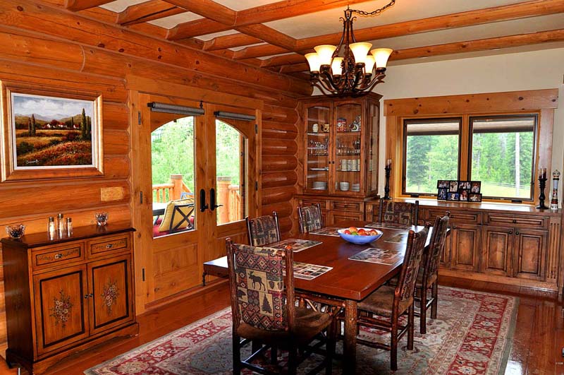 A cozy dining room with wooden walls and furniture, featuring a large table surrounded by chairs, a chandelier overhead, and large windows offering views of greenery outside. There are decorative items on shelves and a painting on the wall.