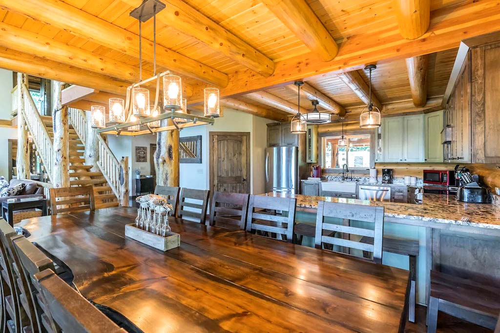 A spacious log home kitchen and dining area featuring a large wooden dining table, exposed wooden beams on the ceiling, and modern appliances in a cozy log cabin style.