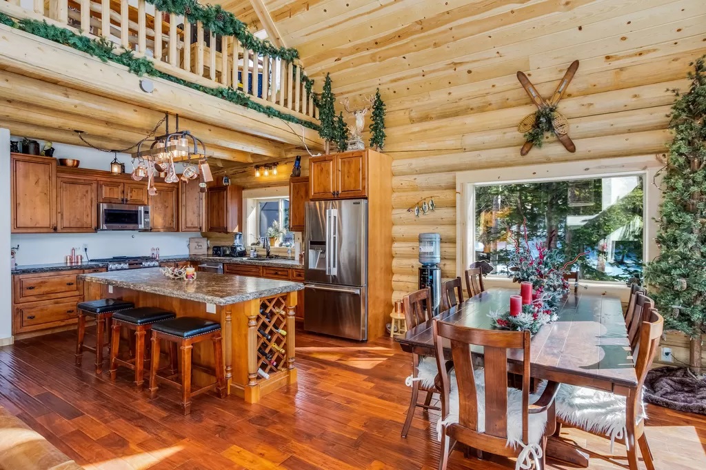 A spacious log home kitchen with wooden cabinets, a large island, and a dining table. The room features high ceilings with exposed beams, and decorations like garlands and holiday accents are present.