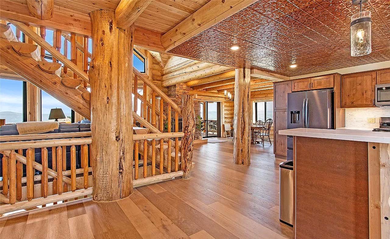 A spacious interior of a log home featuring wooden beams, a staircase, and an open kitchen area with modern appliances and natural light coming through large windows.