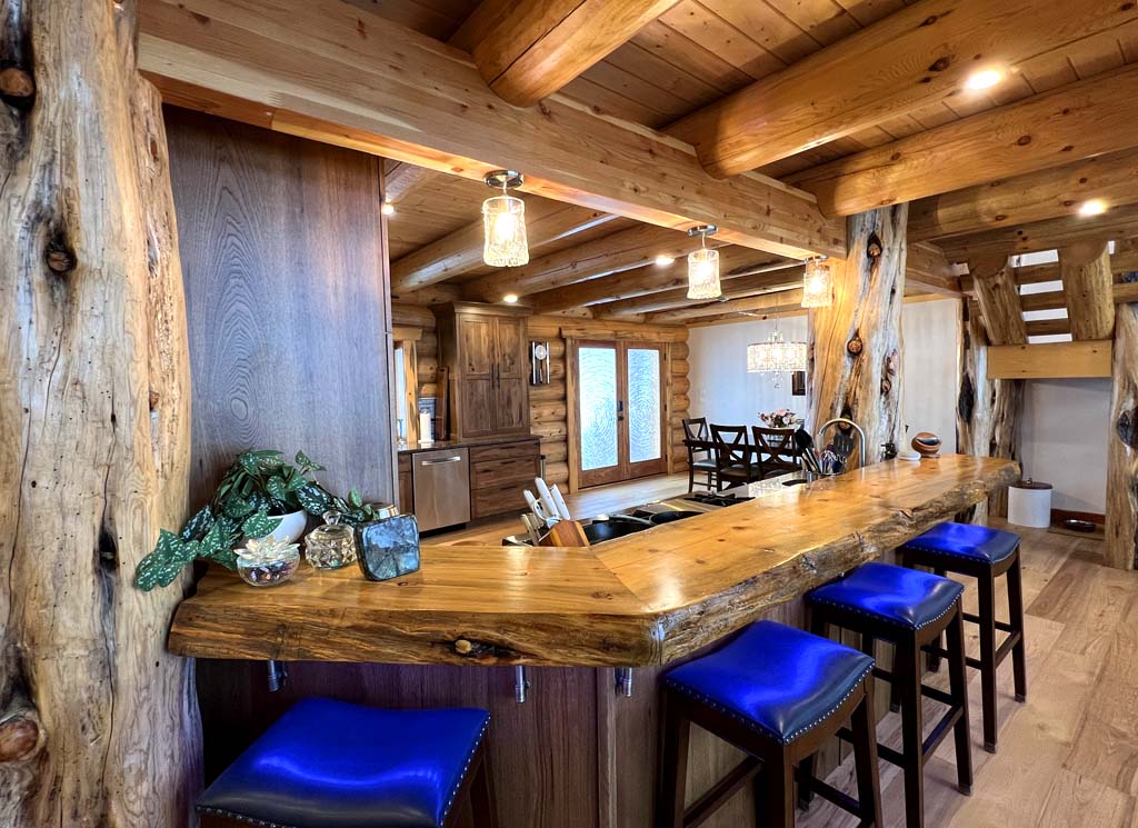 A rustic log home kitchen bar area with a wooden countertop and blue upholstered stools. The bar features pendant lighting and is surrounded by log cabin-style wood elements. There’s an open layout leading to a dining space, with large windows allowing for natural light.