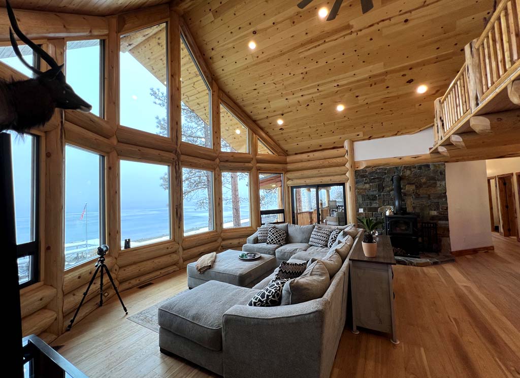 A cozy log home interior featuring large windows, a comfortable sectional sofa with various cushions, wooden construction elements, and a view of the outdoors. A tripod is positioned nearby, and there's a stone fireplace in the background.