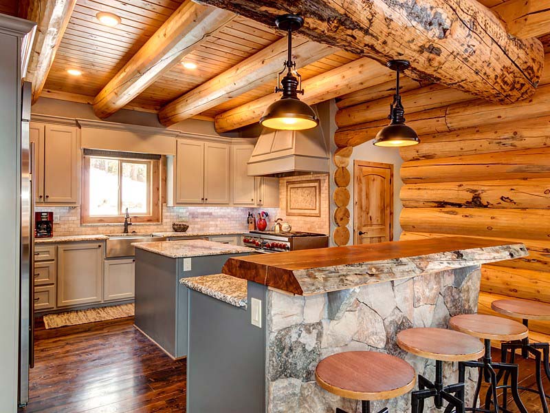 A rustic log home kitchen featuring log cabin elements, wooden beams on the ceiling, a large stone countertop island with bar stools, and modern appliances.
