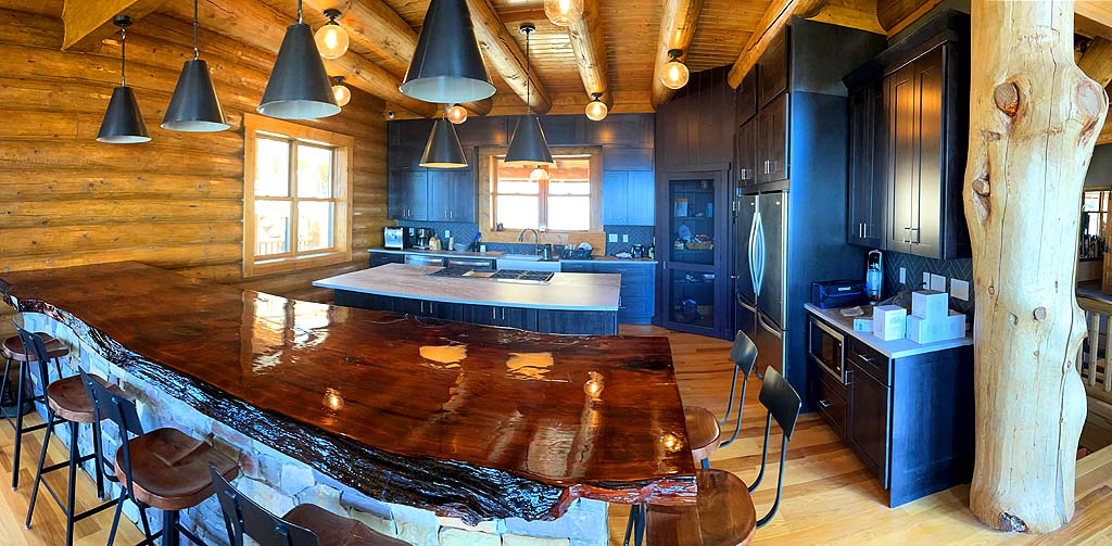 A modern log home kitchen with a wooden log design, featuring dark cabinets, a large island with a glossy countertop, pendant lighting, and stainless steel appliances. The space is bright due to large windows and an open layout.