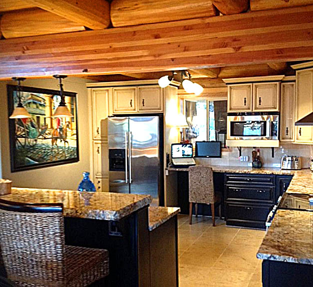 A spacious log home kitchen with wooden beams on the ceiling, featuring granite countertops, a large refrigerator, microwave, and a dining bar with woven chairs. There is a small desk area and artwork on the walls.
