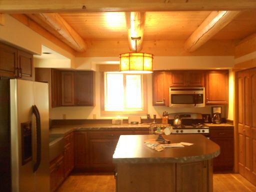A warm, wooden log home kitchen featuring dark cabinets, stainless steel appliances, and a central island with a countertop. Natural light enters through a window, illuminated by a pendant light above the island.