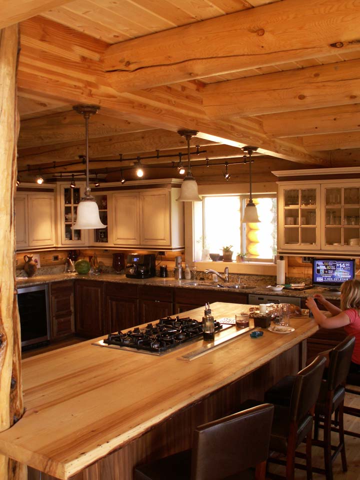A cozy log home kitchen with wooden beams, a large central island featuring a gas stovetop, and wooden cabinets. Natural light comes through a window. There are stools at the island and someone is sitting and using a device.