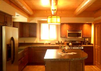 A warm, wooden log home kitchen featuring dark cabinets, stainless steel appliances, and a central island with a countertop. Natural light enters through a window, illuminated by a pendant light above the island.