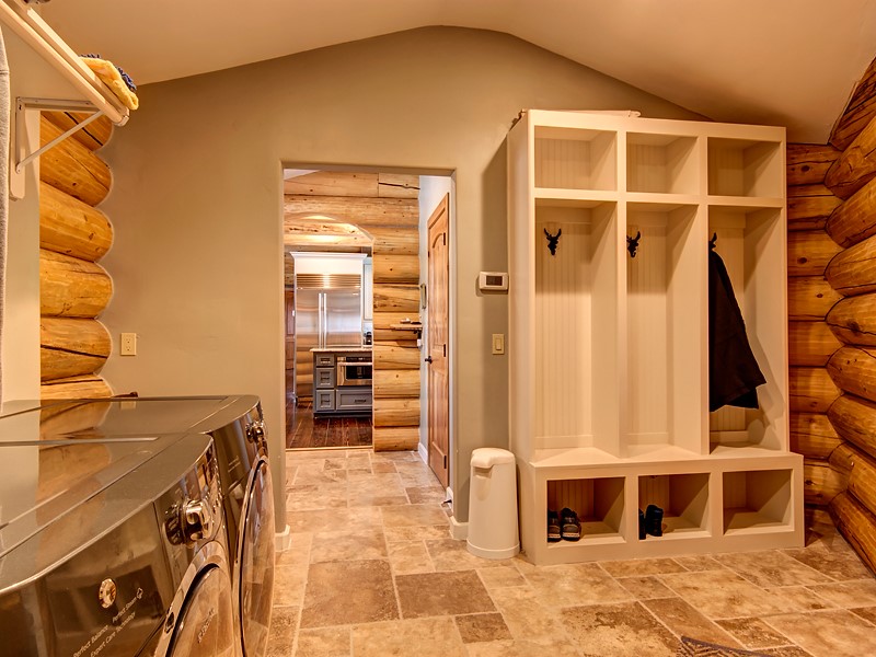A laundry room with a log cabin style, featuring a washer and dryer, a built-in mudroom unit with hooks, and a hallway leading to the kitchen. The flooring is tiled and the walls are painted in neutral colors.