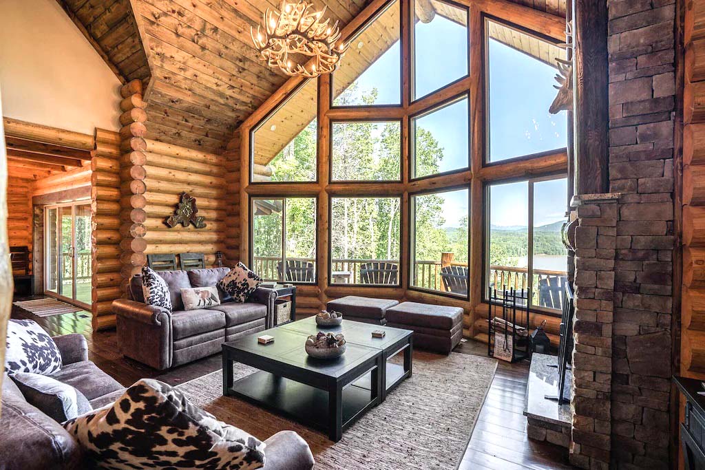 A spacious log home interior featuring large windows with scenic views, a cozy seating area with gray couches and decorative pillows, a coffee table in the center, and a stone fireplace. Sunlight fills the room, highlighting the natural wood details of the walls and ceiling.log home living room area North Carollina rental 1024