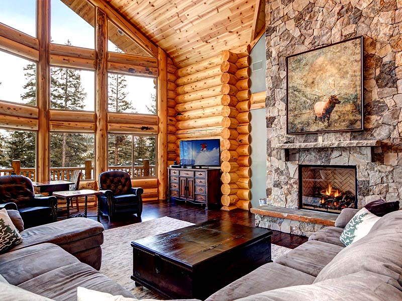 A cozy log home living room featuring a stone fireplace, large windows with forest views, comfortable seating, and wooden accents.