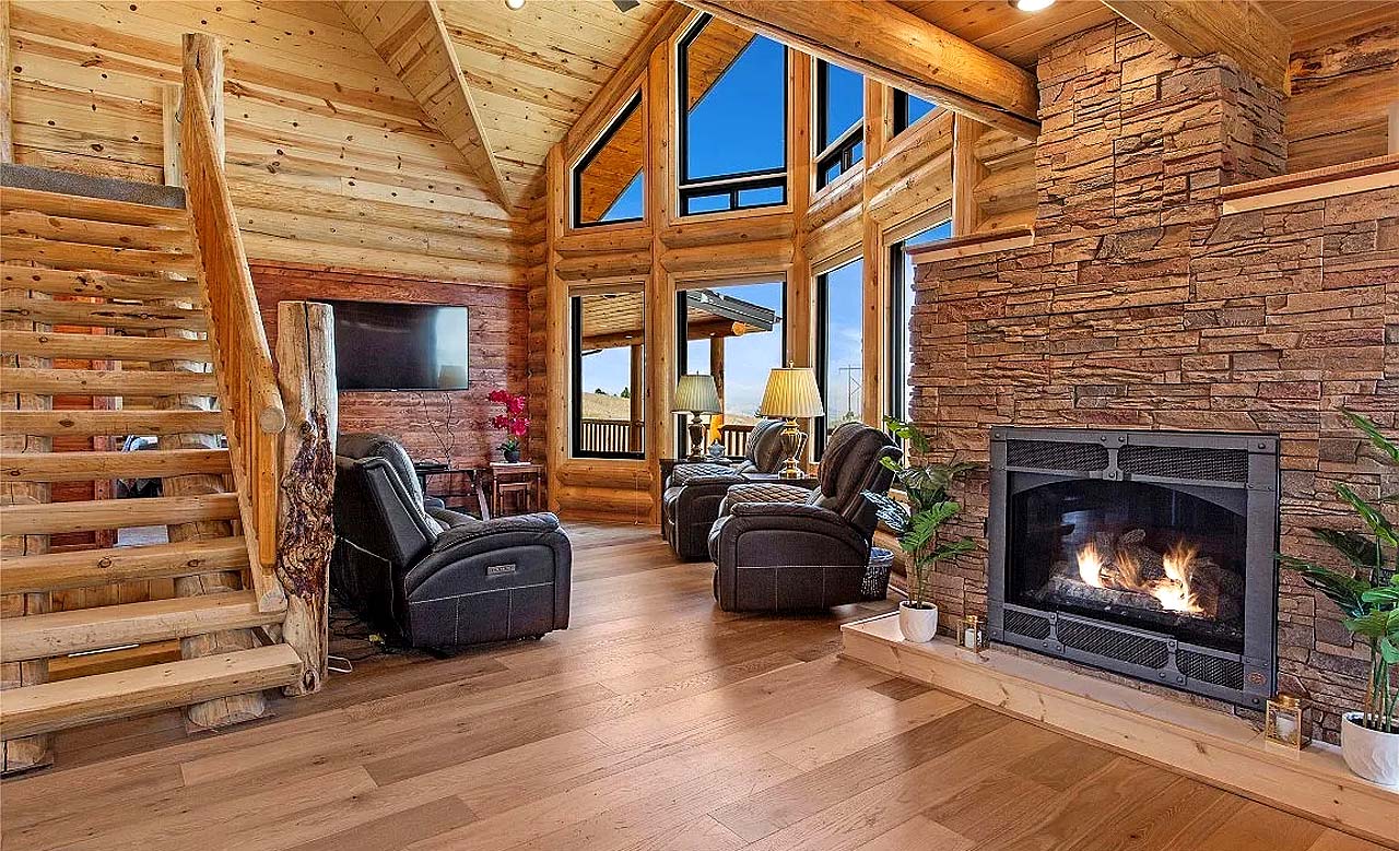 A cozy log home interior featuring a stone fireplace, wooden beams, large windows with a view, leather recliner chairs, and wooden stairs.