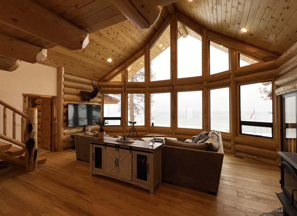 A cozy, log home interior with large windows offering a view of the outdoors. The room features wooden walls and ceiling, a comfortable sofa, a coffee table, and a TV. There is also a staircase leading to another level.