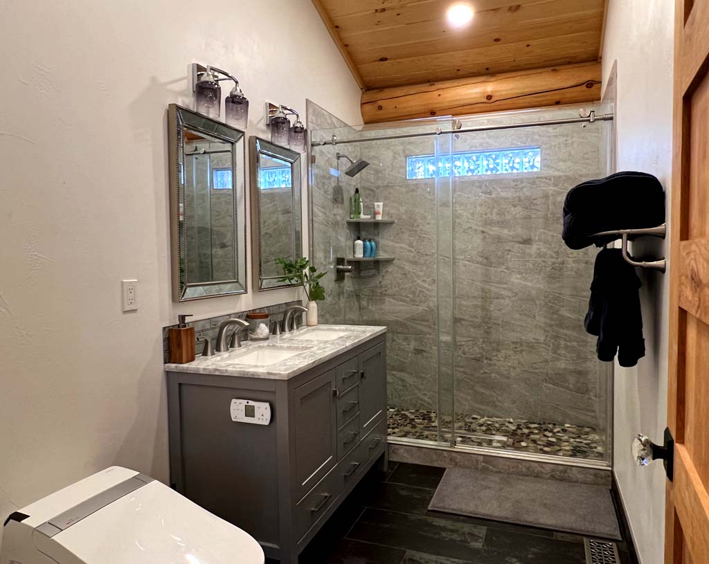 A modern log home bathroom featuring a double vanity with a marble countertop, two mirrors above the sinks, a shower enclosed in glass, and natural wood accents on the ceiling. The space has dark tile flooring and includes toiletries and decorative elements.