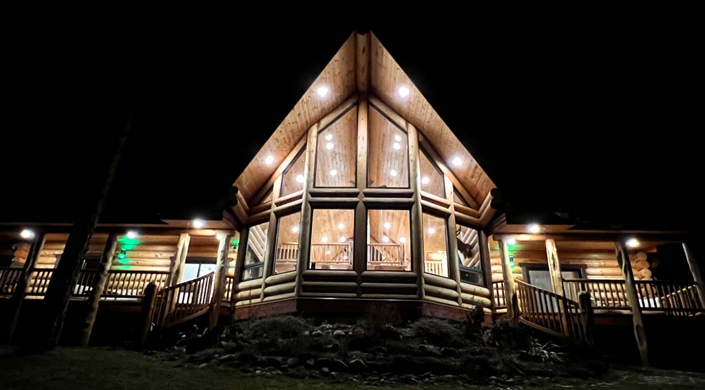 A large log cabin illuminated at night, showcasing a triangular roof, large glass windows, and outdoor wooden decks.