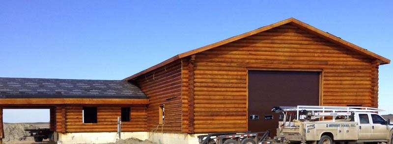 A wooden log cabin-style building with a large garage door on one side, surrounded by a construction area and a pickup truck parked nearby. The sky is clear and blue.
