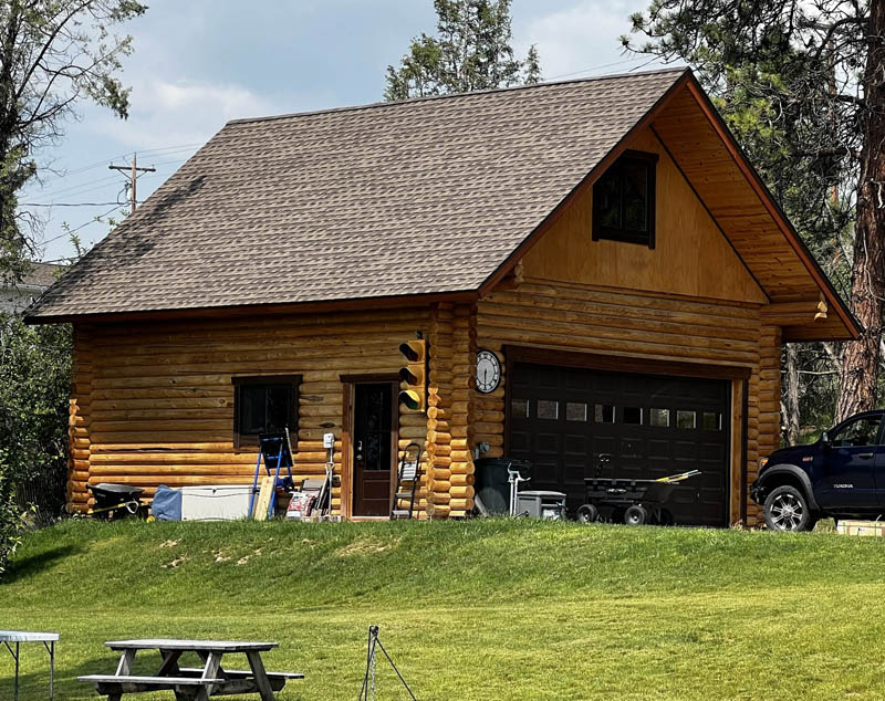 A log cabin-style garage with a brown roof, a large garage door, and several outdoor items and tools around it, set on a green lawn with trees in the background.