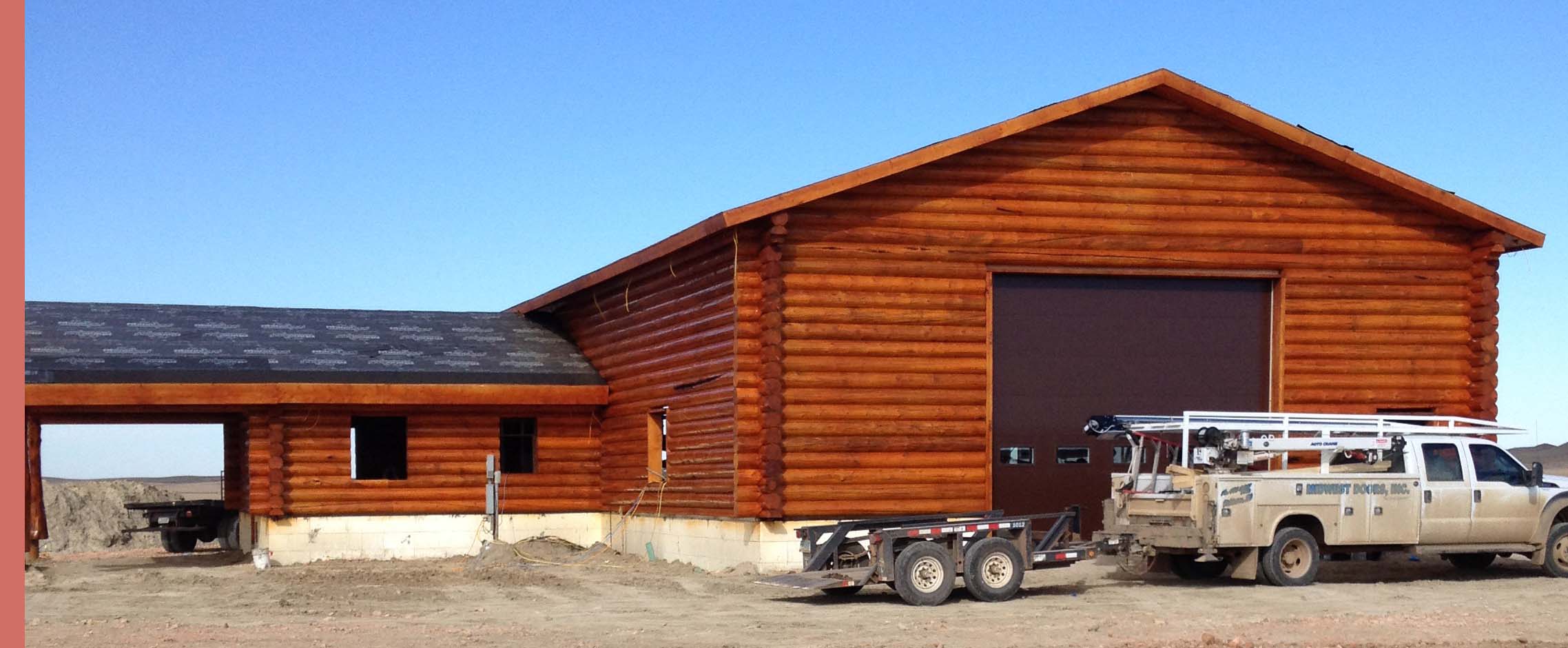A large log cabin structure with a gabled roof and a separate, smaller area attached. A truck and trailer are parked outside.