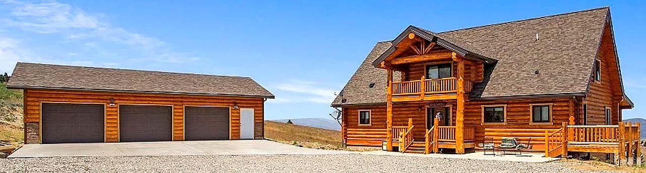 A large log cabin-style house with a front porch, surrounded by open land, and a separate garage with three doors.