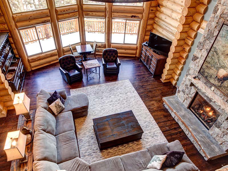 A cozy log home interior featuring a spacious living area with a large sectional sofa, two armchairs, a rustic coffee table, and a stone fireplace. Large windows provide plenty of natural light and showcase the wooded surroundings.