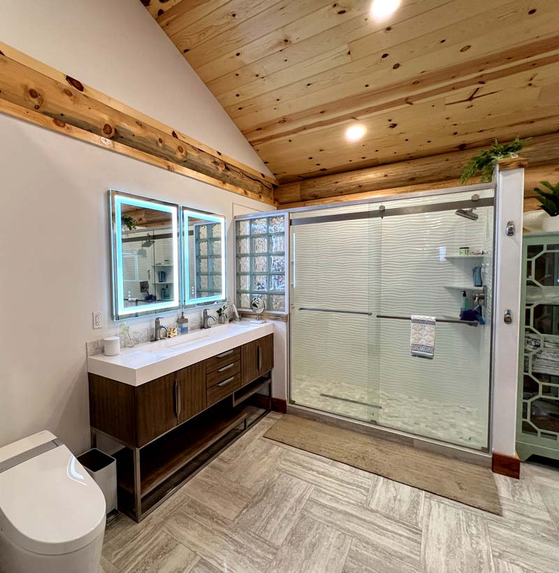 A modern log home bathroom featuring a wooden ceiling, a double vanity with a mirror that has LED lighting, and a spacious glass shower enclosure.