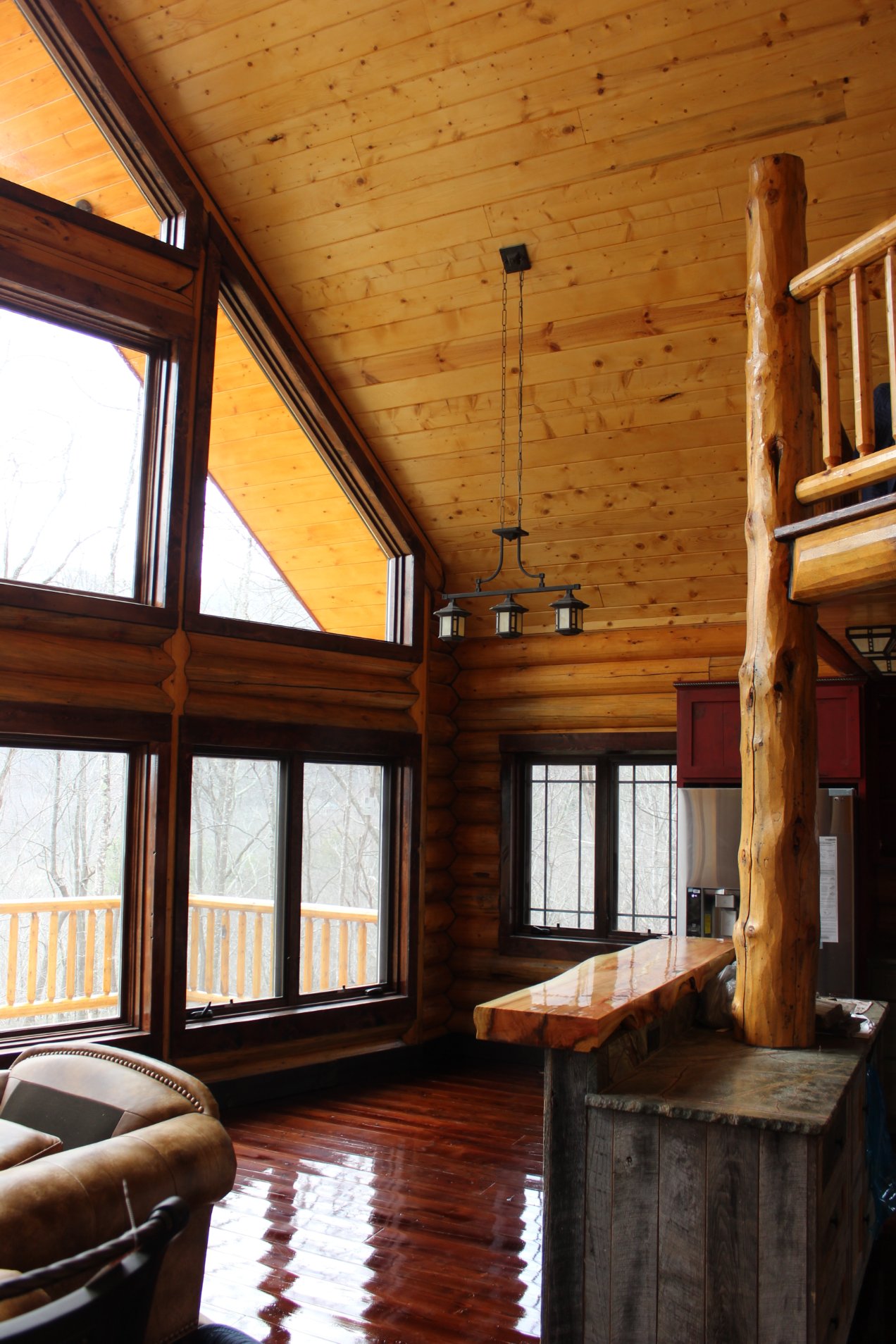 A log home spacious interior of a cabin with wooden walls and ceilings, featuring large windows that provide natural light. There is a wooden bar area and rustic chandeliers hanging from the ceiling.