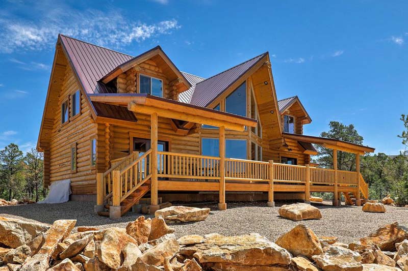 A large log home with a triangular roof, large windows, and a wooden deck surrounded by rocks and trees under a clear blue sky.