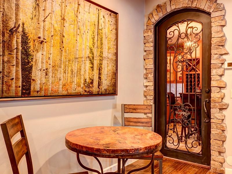 A cozy dining area featuring a round wooden table with two chairs, a large framed artwork depicting trees in autumn, and an ornate iron door with stone accents.
