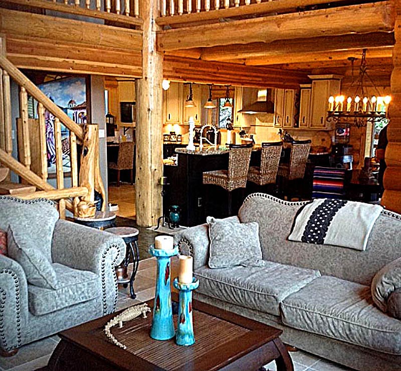 A cozy log home living room with log cabin features, including wooden beams and a rustic dining area visible in the background. There are two plush sofas, a coffee table with decorative items, and an inviting ambiance.