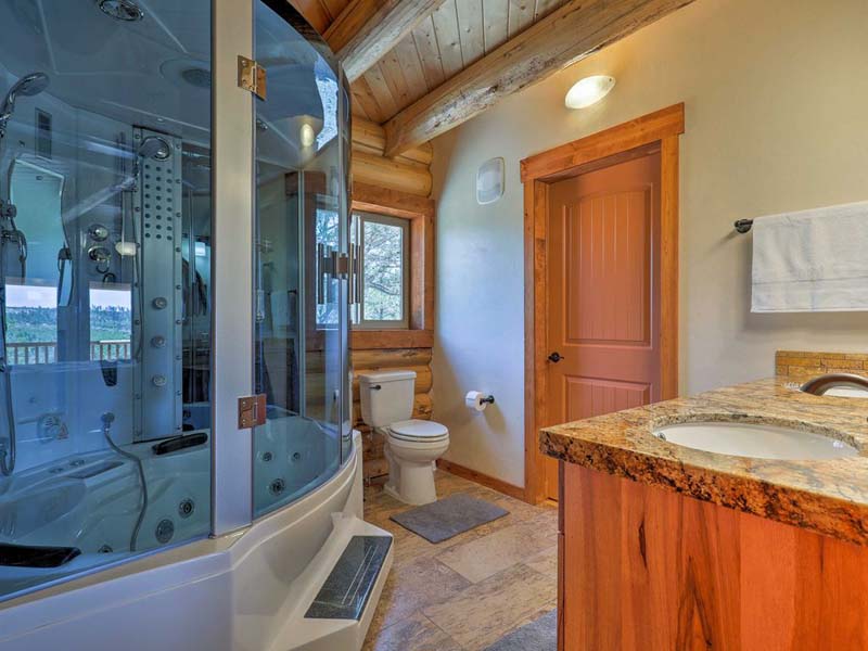 A modern log home bathroom featuring a glass shower, toilet, and sink with granite countertop. The walls are wooden, and there's a large window providing natural light.