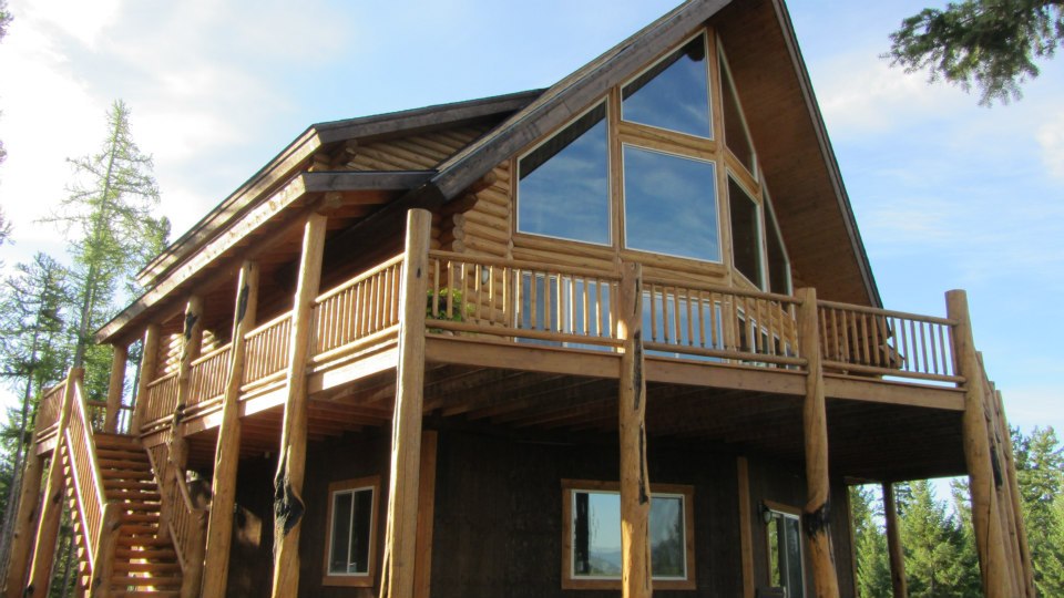 A wooden log home with large windows and a sloped roof, elevated on stilts with a staircase leading to the entrance. Surrounded by trees and blue sky.